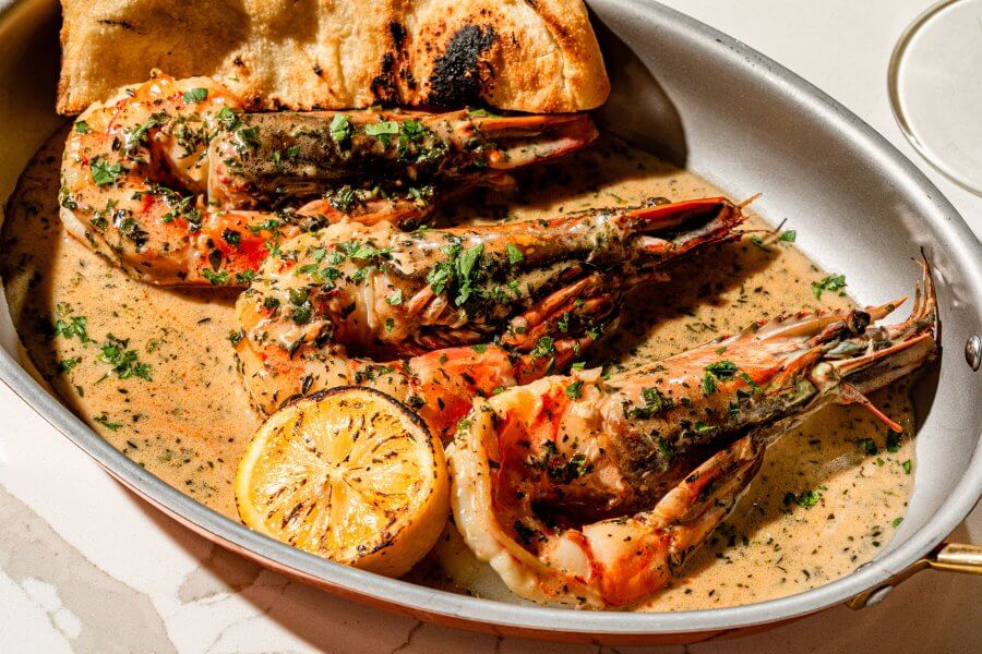 Grilled shrimp with herbs is elegantly served in a metal dish with creamy sauce, a charred lemon half, and toasted bread—a must-try at one of Houston's top new restaurants.