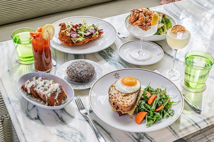 An exquisite spread on a marble table awaits, featuring salad, shrimp cocktail, croque monsieur with greens, a rich chocolate dessert, seasoned fries, and assorted drinks. 