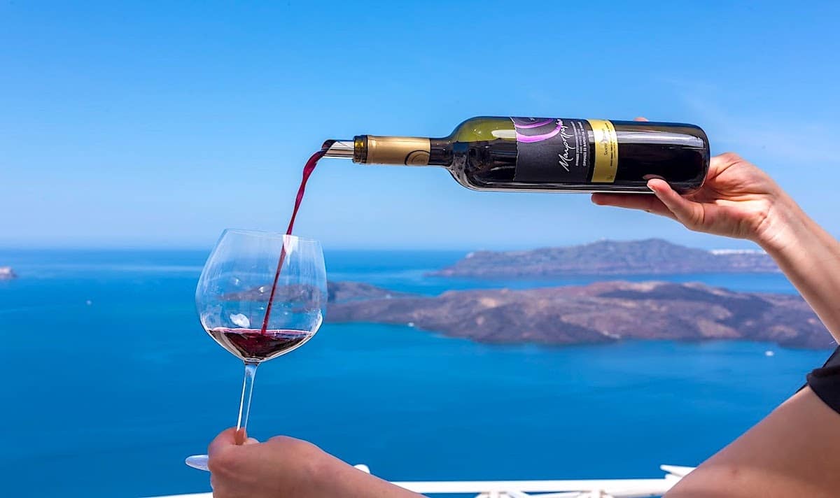 Image of someone pouring red wine into a wine glass with the blue Greek sea in the background.