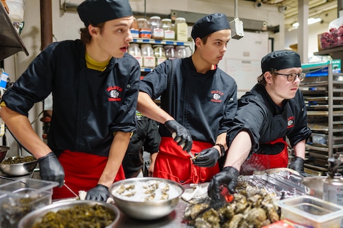 Three students watch and work as they learn to shuck oysters