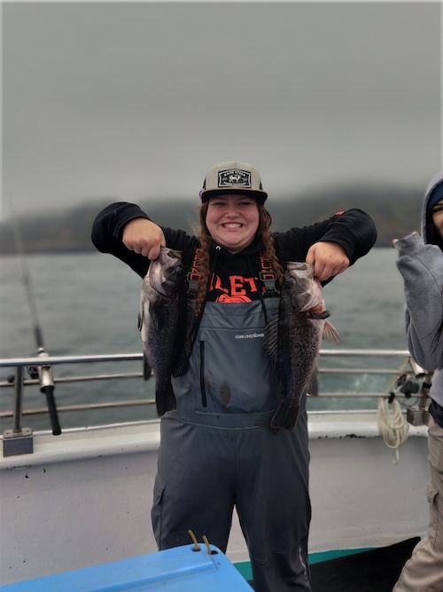 Zoey is shown on a fishing boat posing for a photo as she holds up two black rockfish by the mouth
