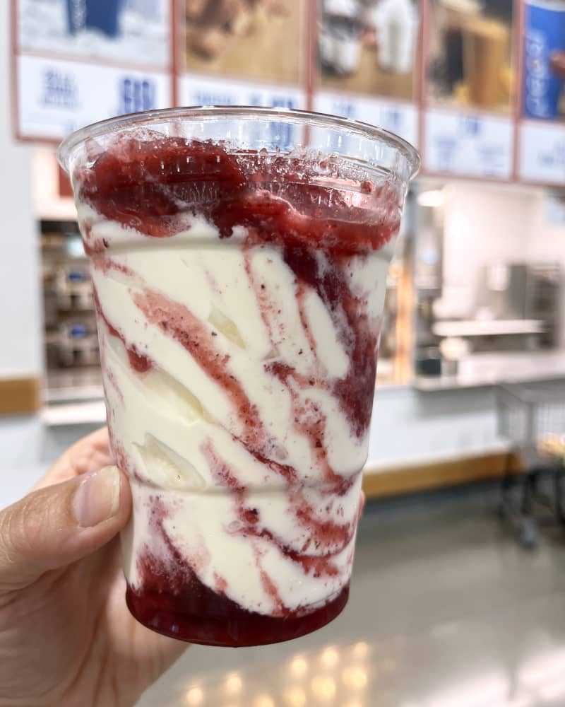 Someone holding up a Costco strawberry ice cream sundae in a plastic cup in the food court