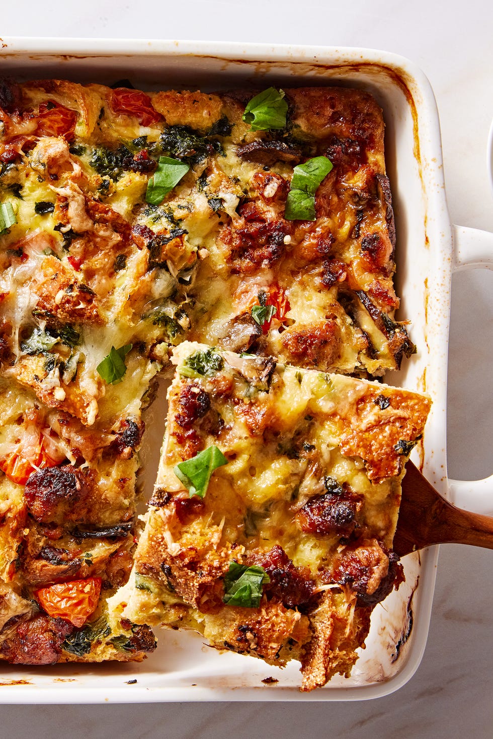 eggy breakfast strata featuring sausage, tomatoes, bread, and mushrooms, and topped with basil