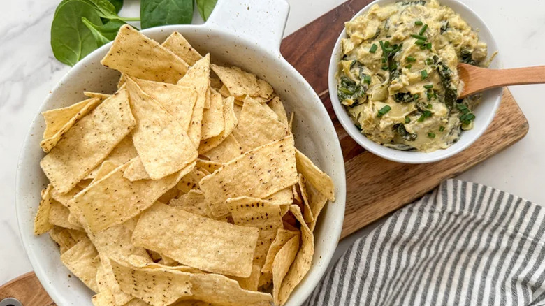 Bowl of chips alongside spinach and artichoke dip on wooden board
