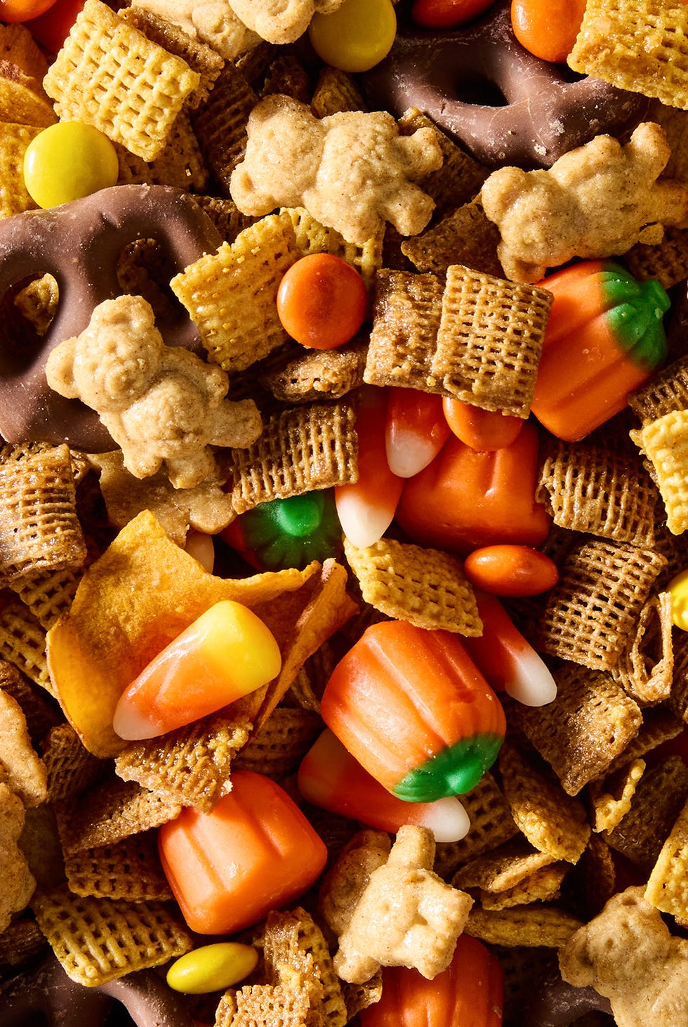 a bowl of halloween themed chex mix with chex cereal, chocolate pretzels, reese's pieces, candy corn, and sweet potato chips