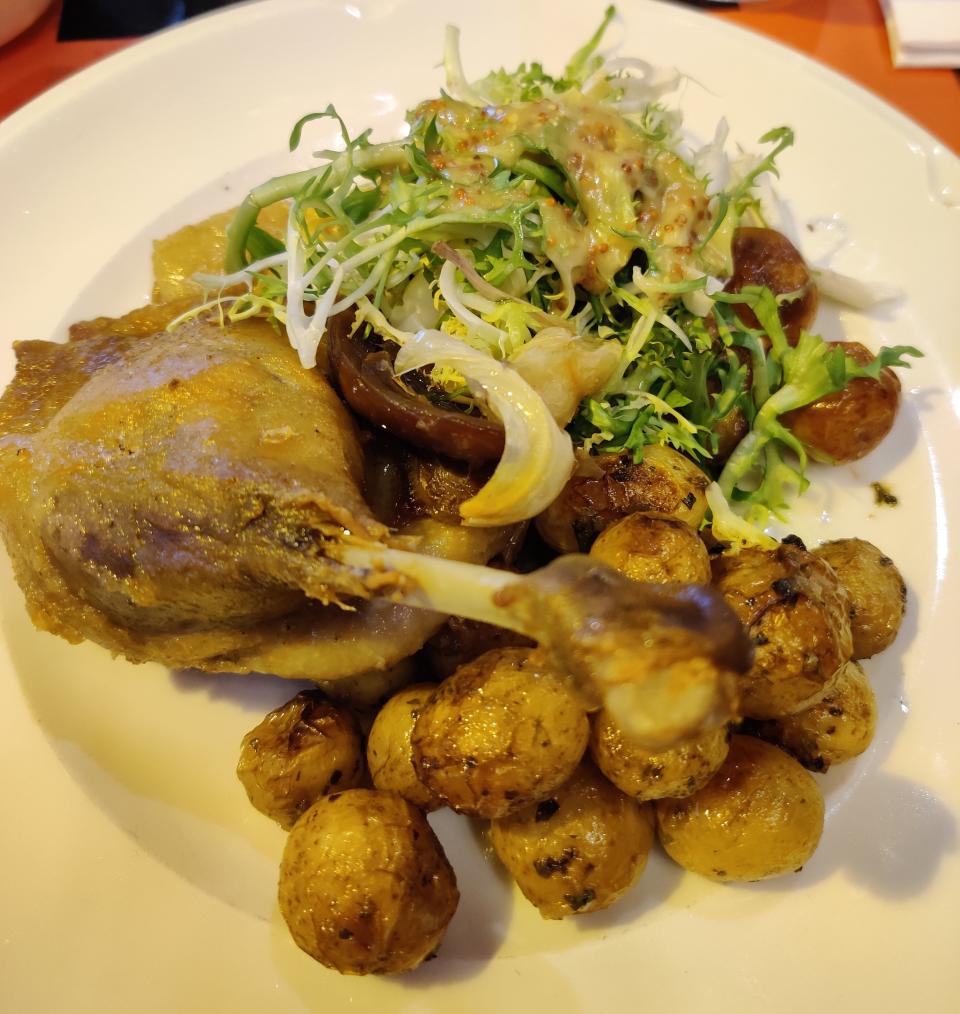 A plate of duck confit served with crispy baby potatoes, frisée salad, grilled mushrooms, and drizzled with a mustard dressing