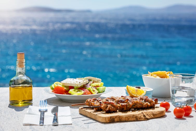 A bottle of olive oil and healthy food on a table, with the sea in the background
