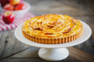 Close-up of baked apple tart on dessert stand with bowl of fresh apples resting on napkin in background.