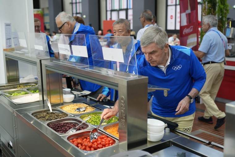 IOC President Thomas Bach tries food from a salad bar while touring the Olympic Village