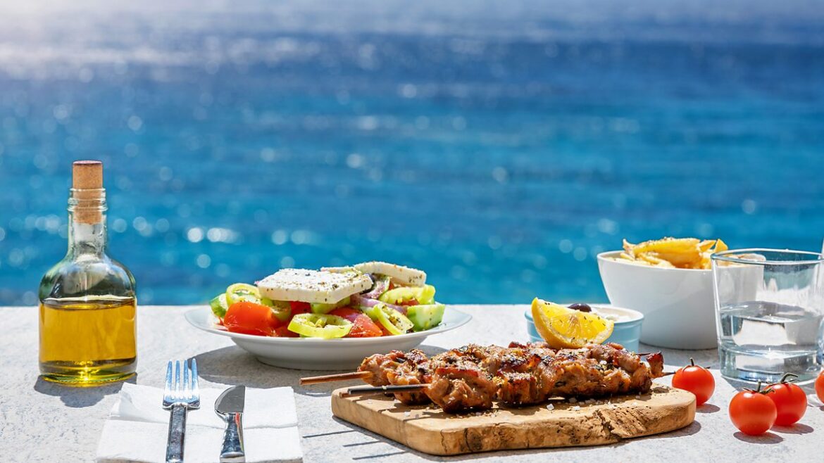 Greek food including kebabs and Greek salad on a table with a bottle of olive oil with the sea in the background