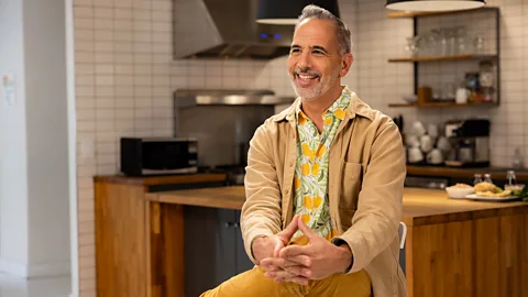 Natassja Ebert Chef Yotam Ottolenghi sits cross-legged on a stool wearing a floral-patterned shirt. Behind him is a soft-focus kitchen backdrop with steel fixtures and wooden accents (Credit: Natassja Ebert)