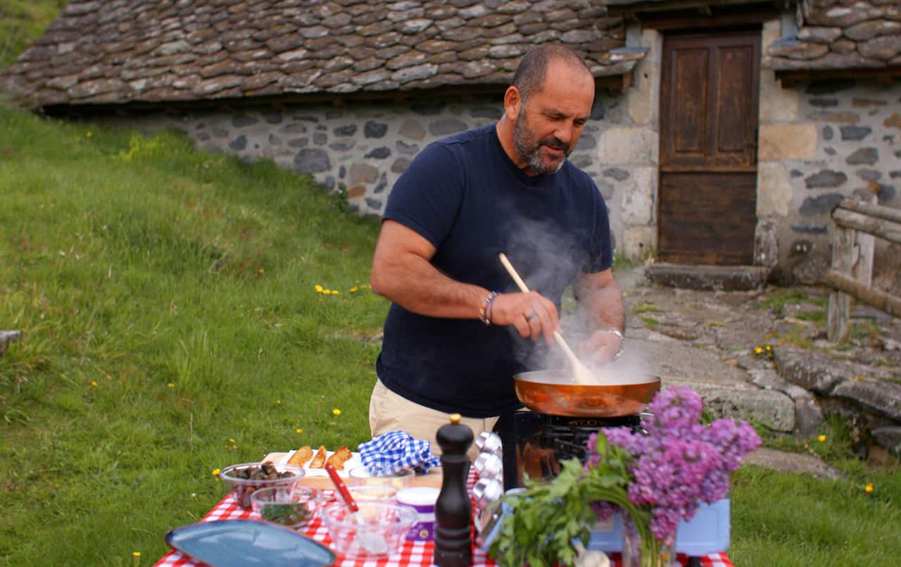A man in a black shirt stands outdoors cooking in front of a stone building. The table in front of him is colourful, with a red checked cloth and a bunch of purple flowers.