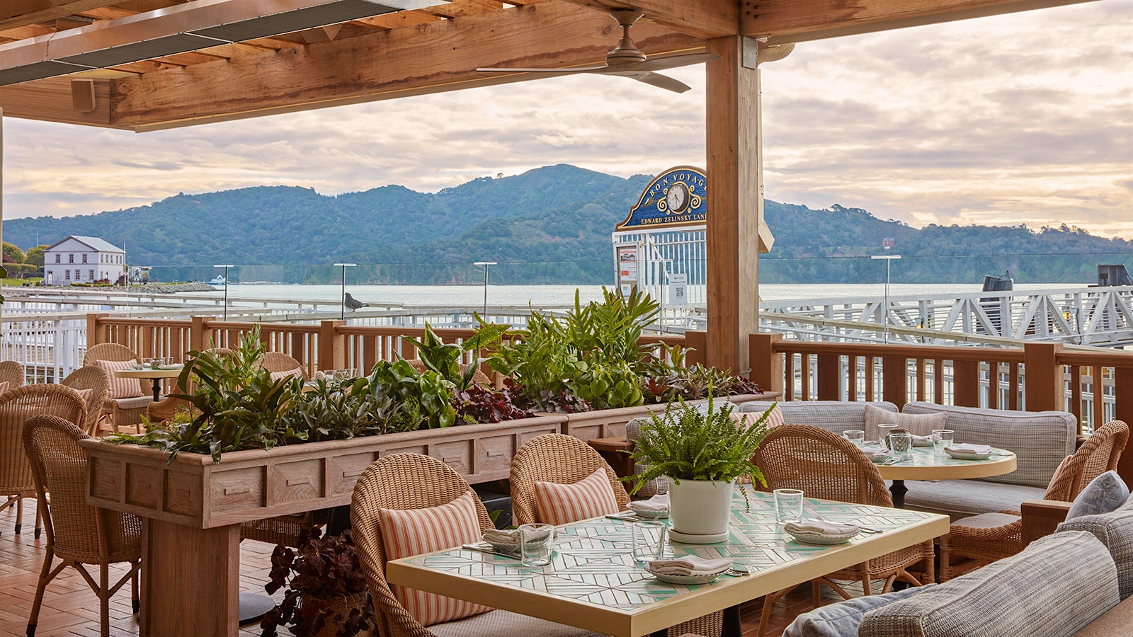 Tile-top tables and wicker chairs outdoors on a deck at the Bungalow Kitchen by Michael Mina, with seaside views of water and forested hills