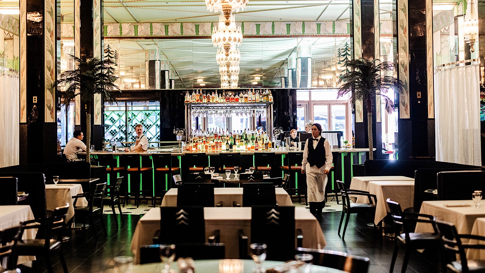Dark wood chairs, tables with white tablecloths, the main bar and restaurant staff in the dining room of Fhima's Minneapolis, with chandeliers hanging overhead