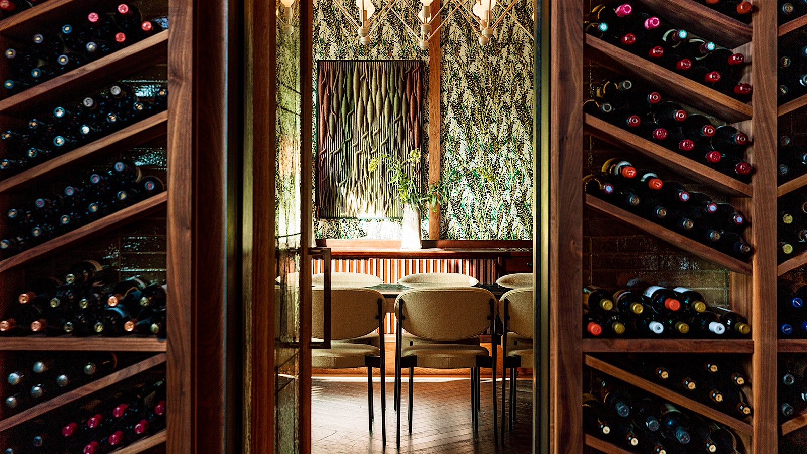 A table and racks of wine bottles at the Peacock Mediterranean Grill, with tan seats, red and yellow flowers in vases and wallpaper depicting green plants