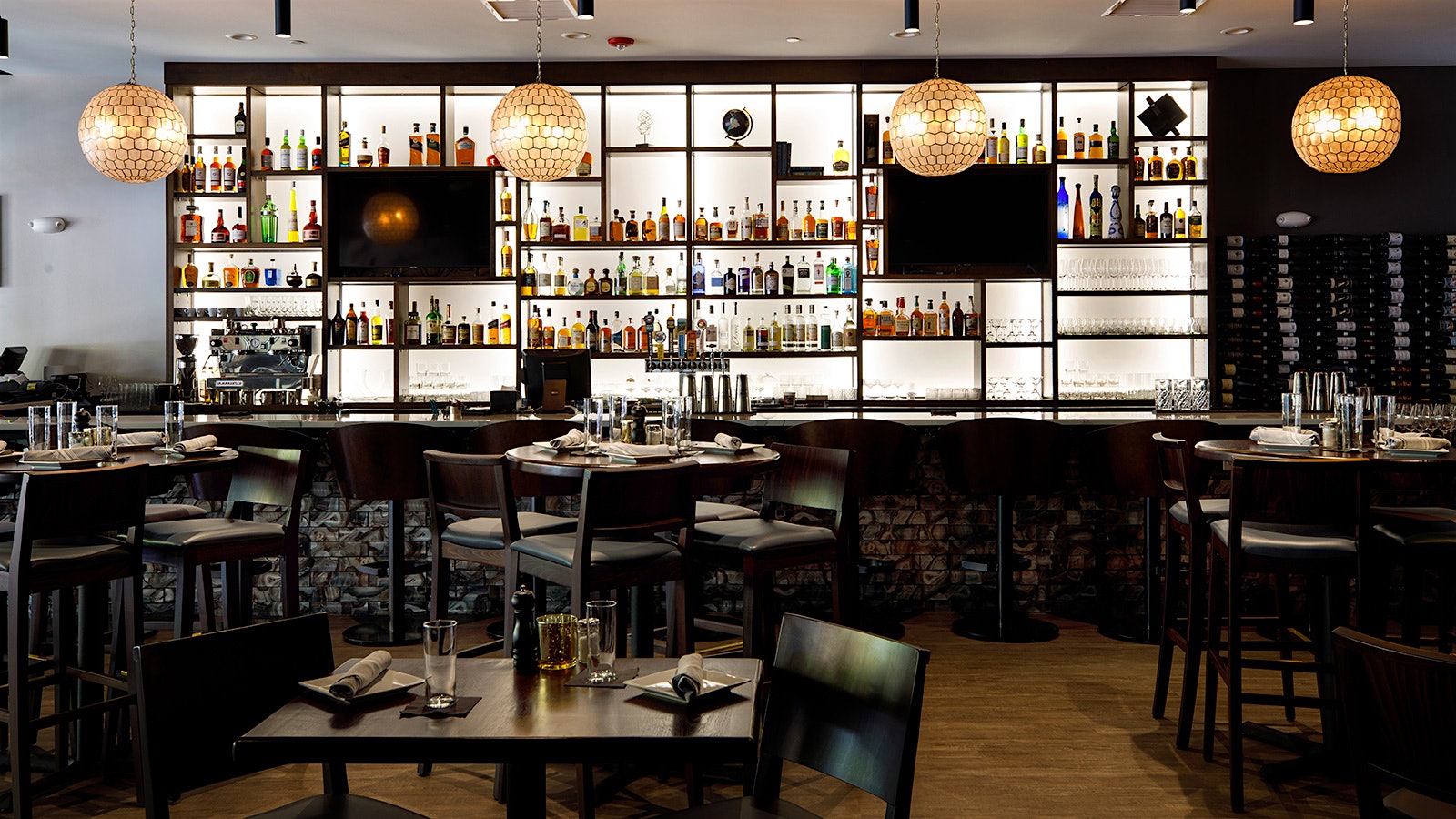 Set wood tables and seats with black leather backing at Wine Dive and Kitchen Lawrence, where there are spherical lighting fixtures and a black-colored bar with lit white backing
