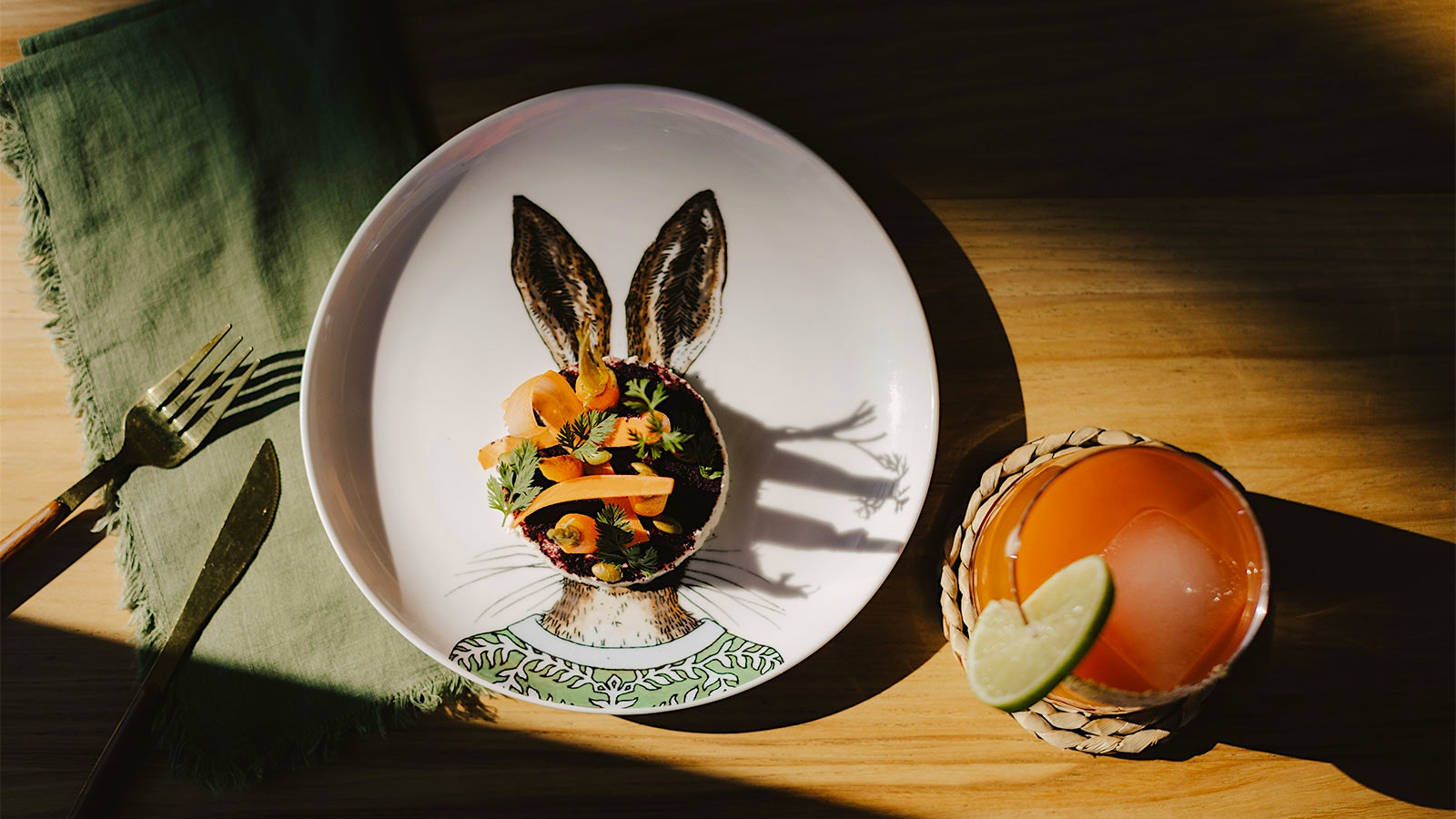 A carrot-based dish served on a plate featuring an illustration of an anthropomorphic rabbit, next to an orange cocktail, at In Bloom