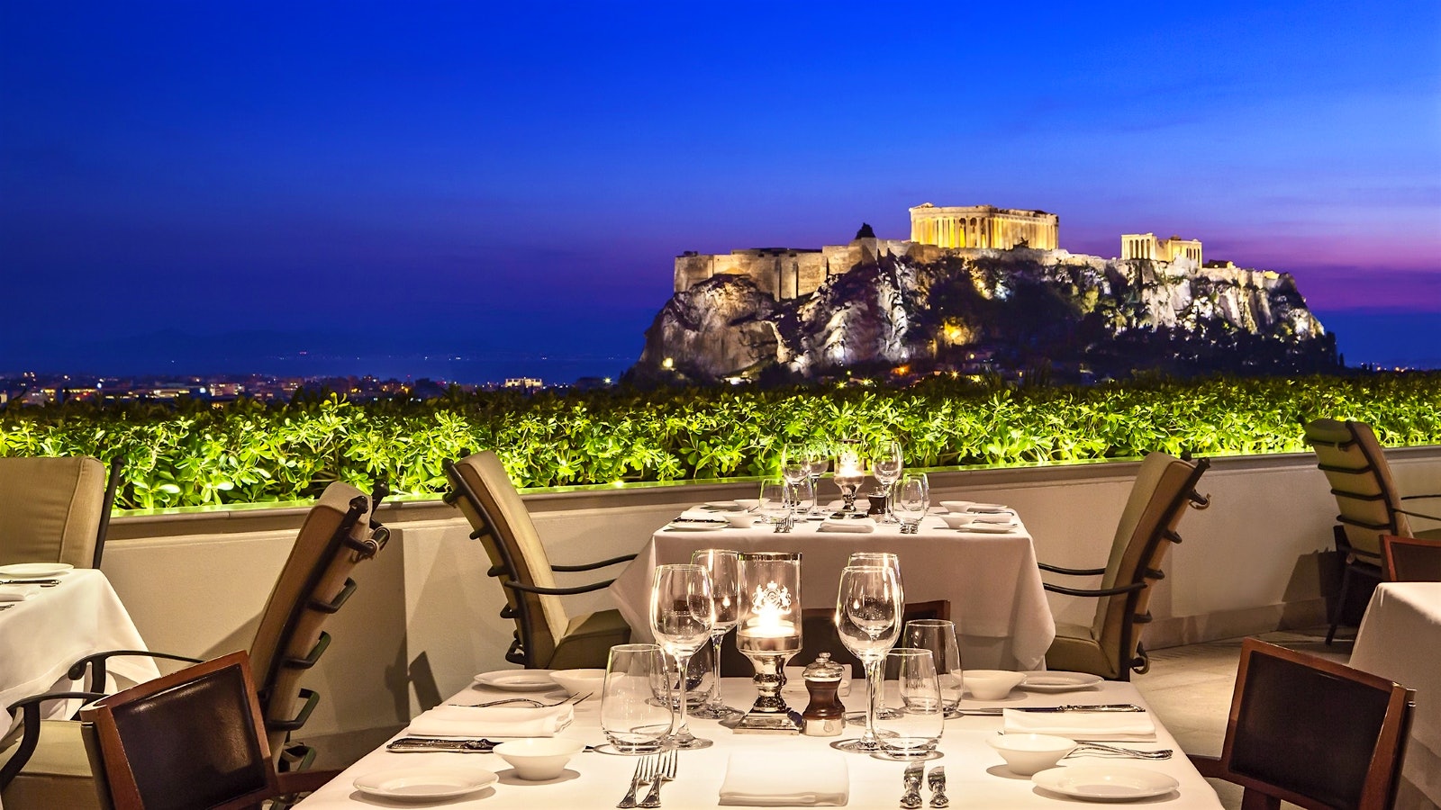  Tables set on the veranda of the GB Roof Garden Restaurant at night, with a view of the ancient Acropolis landmark