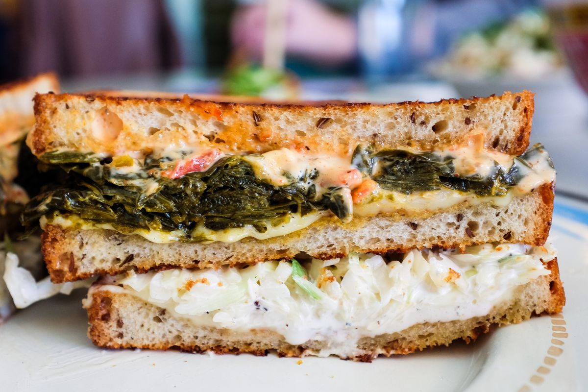 A sandwich of rye bread, coleslaw, collard greens, and sauce is cut in half on a white plate.