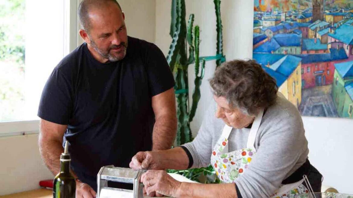 A man in a black t-shirt stands in a home kitchen beside an older woman in an apron, who is working a pasta rolling machine.