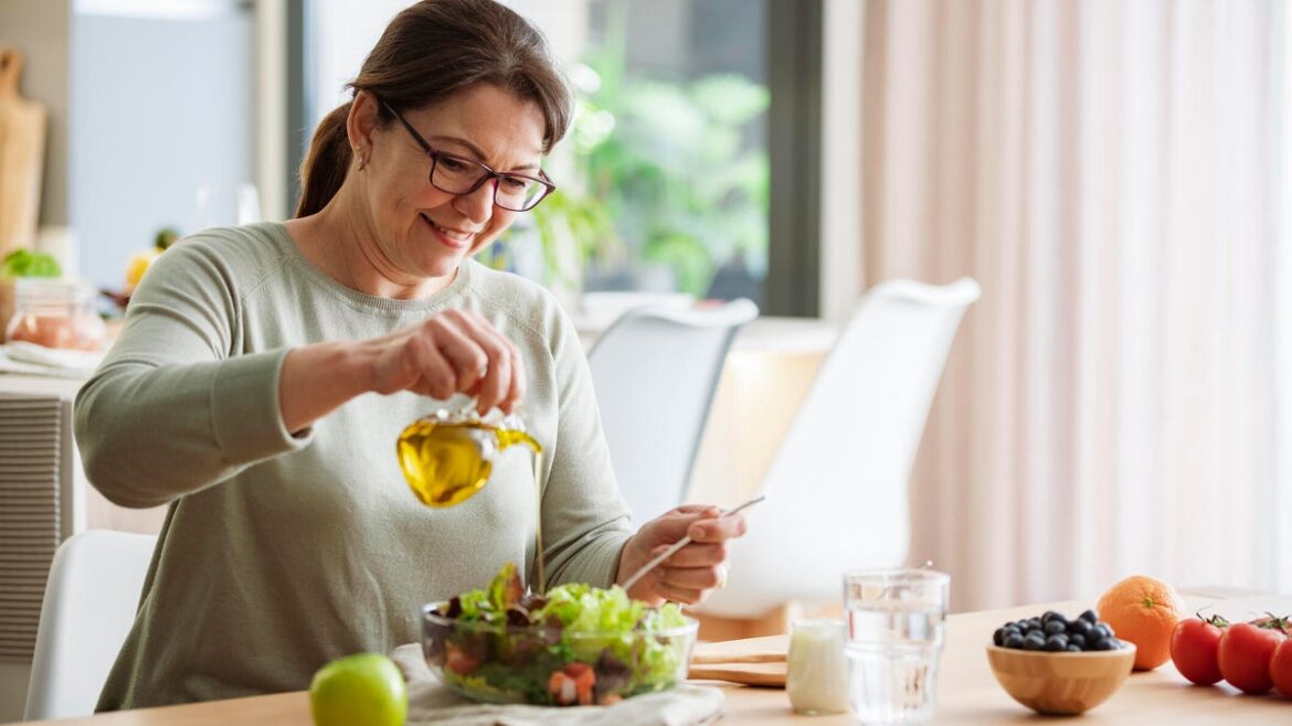 A woman with brown hair in a ponytail and glasses pouring olive oil onto a fresh salad.
