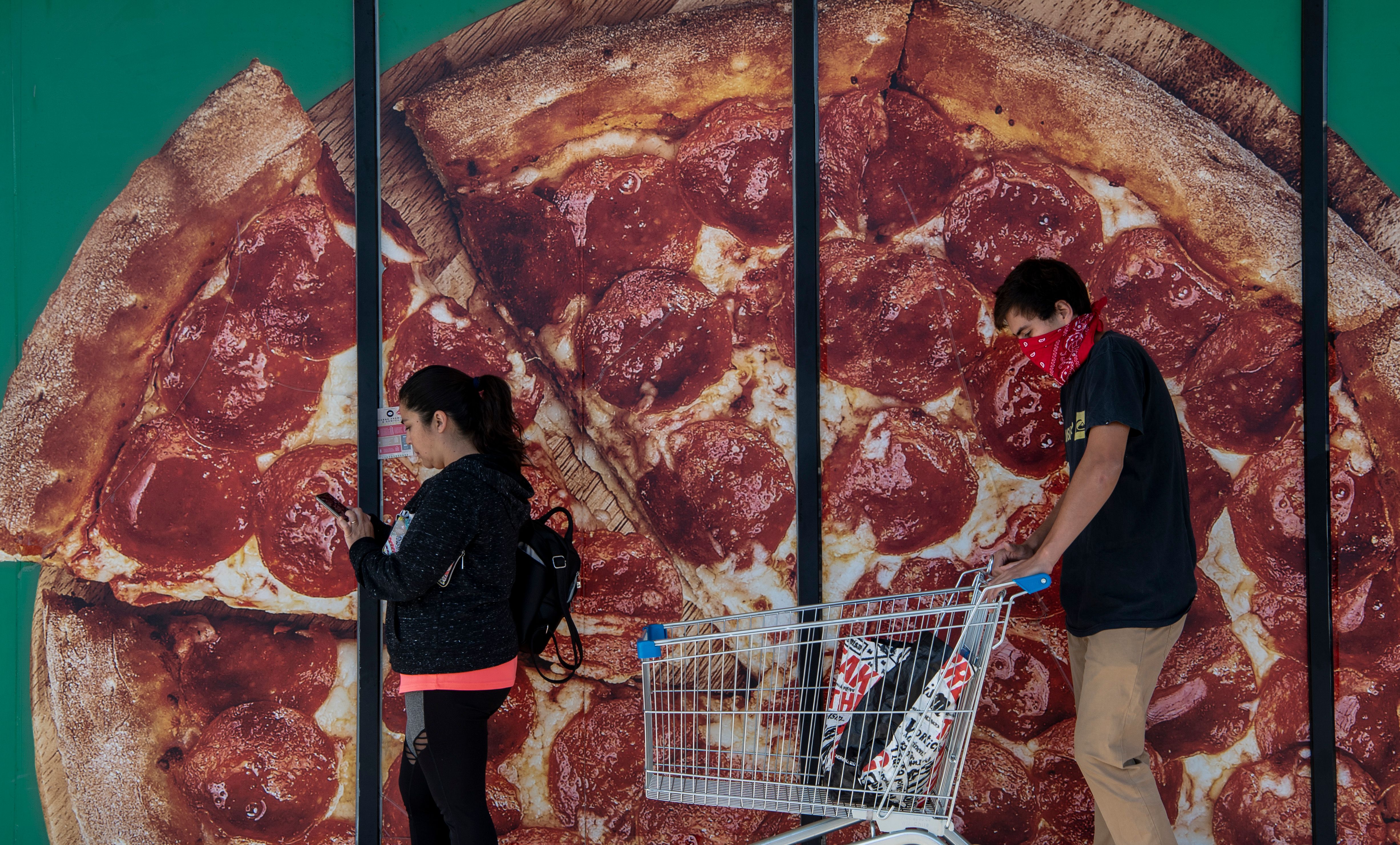 Two people wearing face masks shop at a supermarket with a large pizza advertisement in the background.