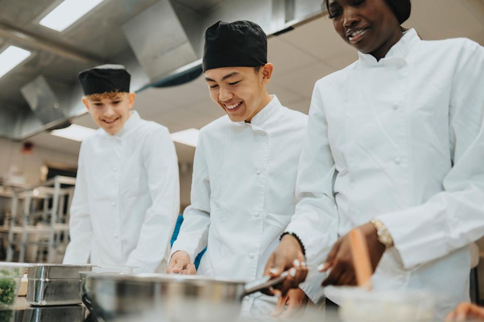 Three chefs in a kitchen, all wearing white uniforms and black hats, focused on cooking together with smiles