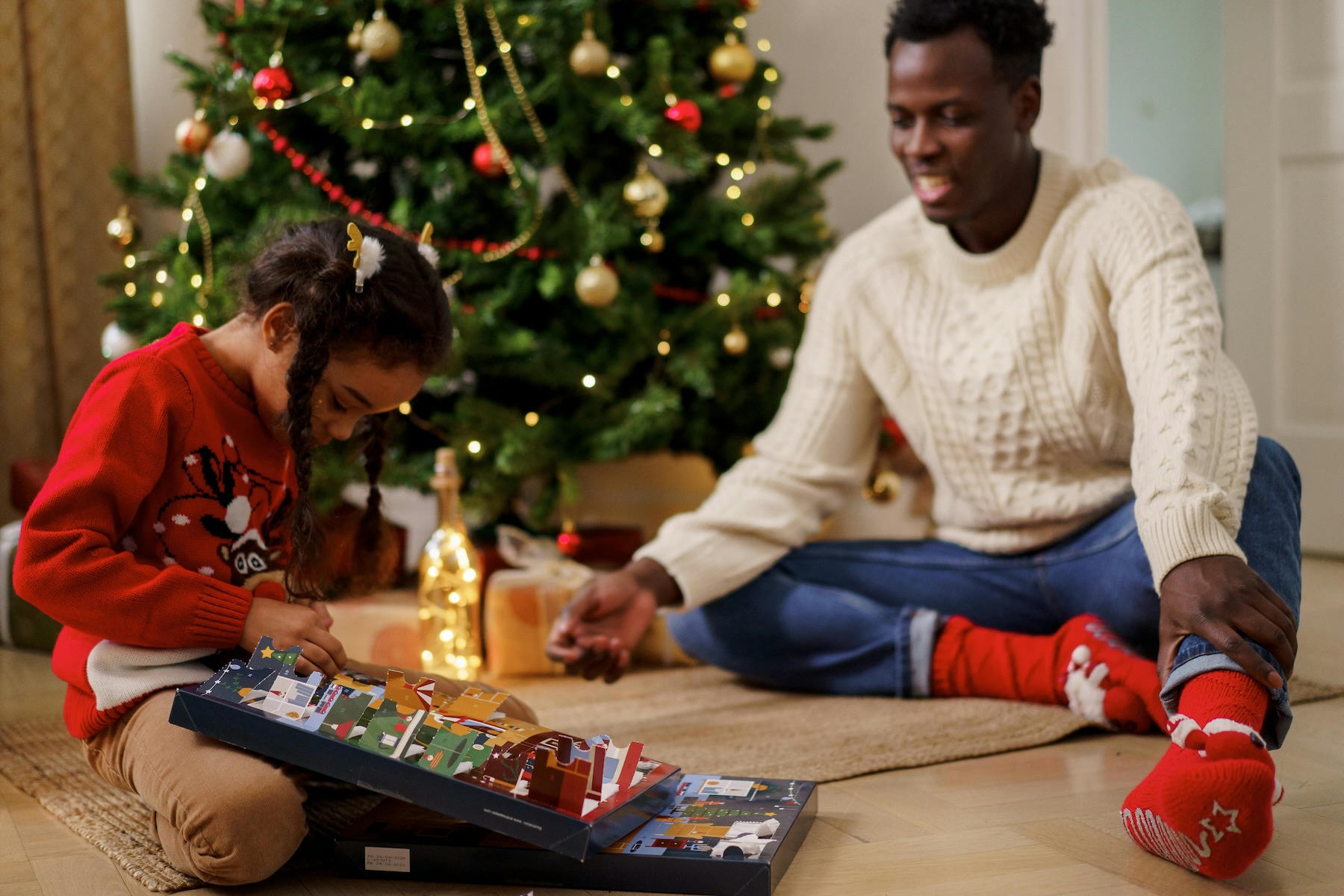 A father and daughter sit near the Christmas tree and open an advent calendar