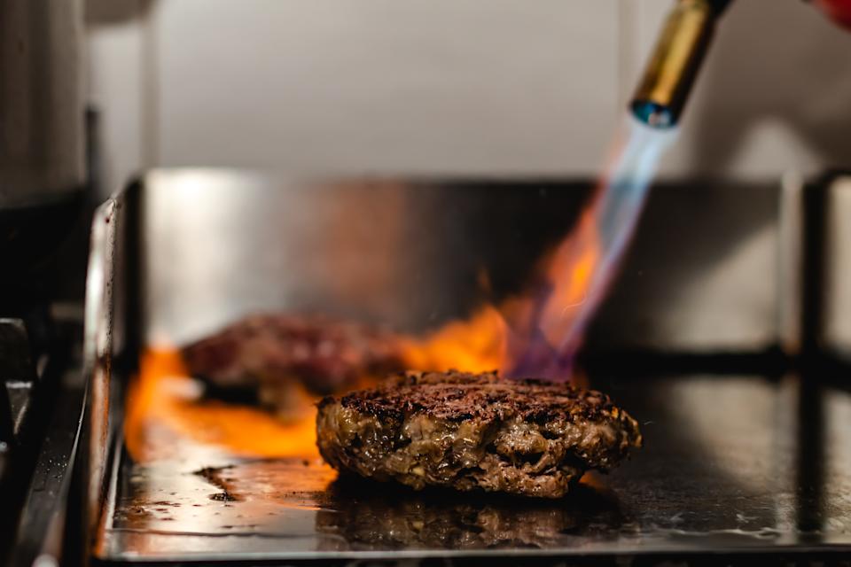 Burger patty on a grill being seared with a flame torch, surrounded by small flames