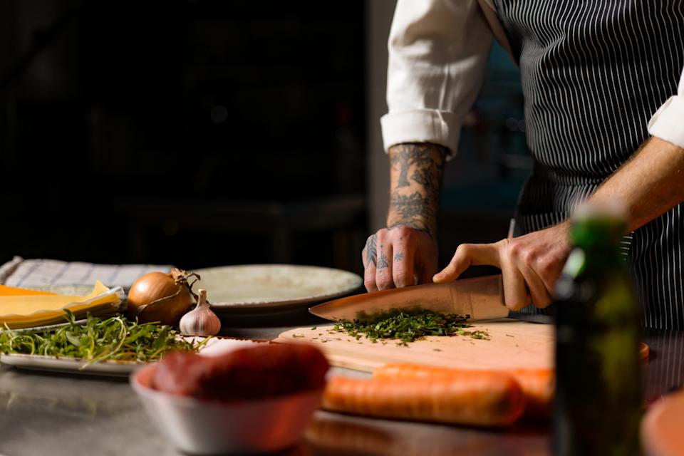 Chef with tattoos chopping herbs on a wooden board in a kitchen, surrounded by fresh ingredients like carrots, garlic, and greens