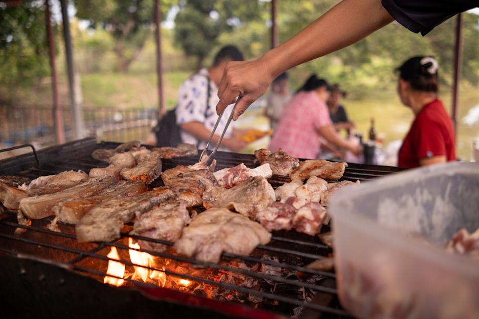 A person grills various meats on an outdoor barbecue, with several people sitting and eating in the background
