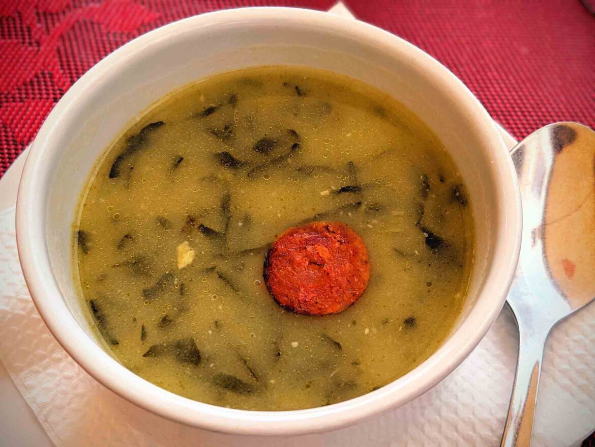 A white bowl filled with a creamy green soup featuring leafy greens and a slice of chorizo in the center. The bowl is placed on a white napkin, and a silver spoon rests on the right side. The tablecloth under the bowl is red with a lace pattern.