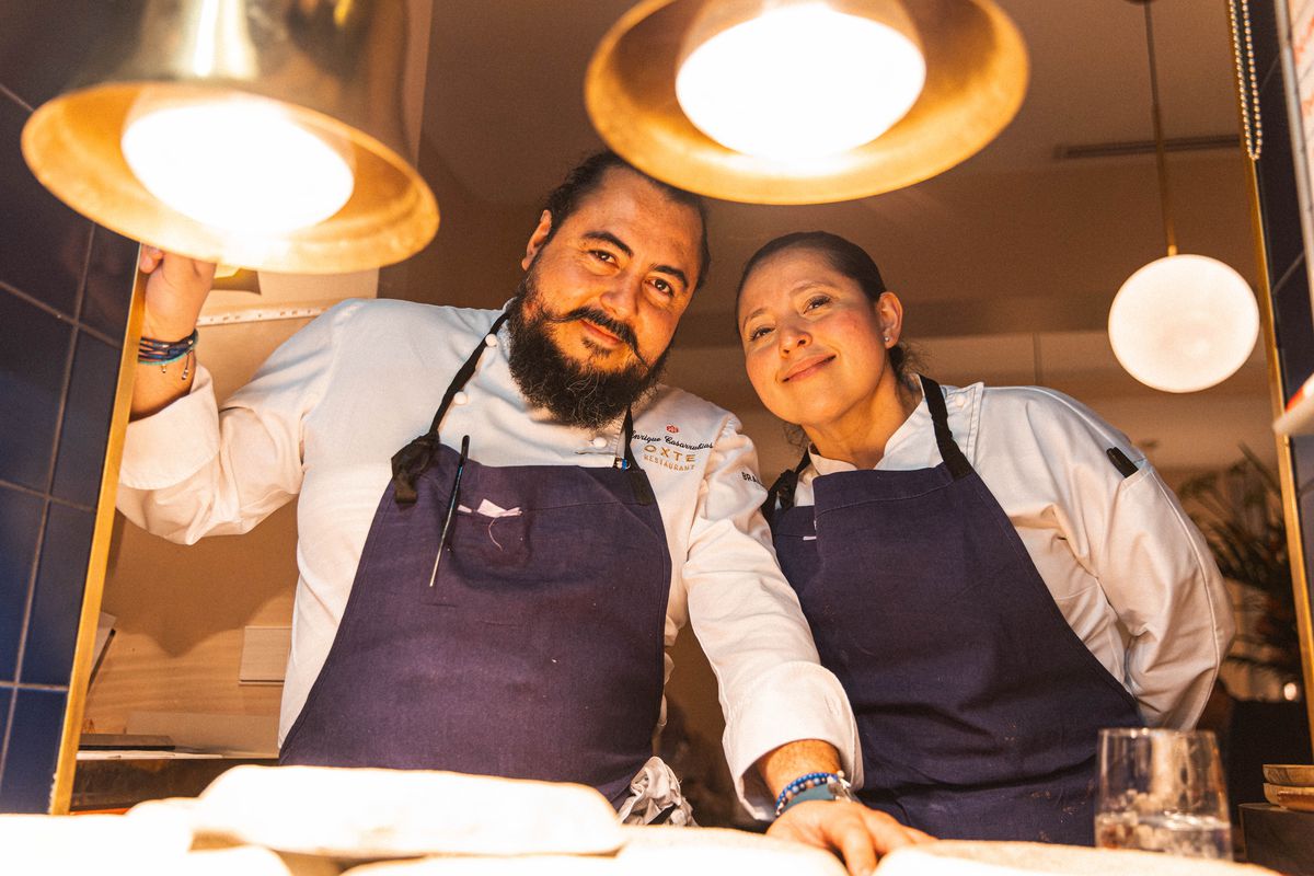 Two chefs look down at the camera, set at the level of dishes on a kitchen counter.
