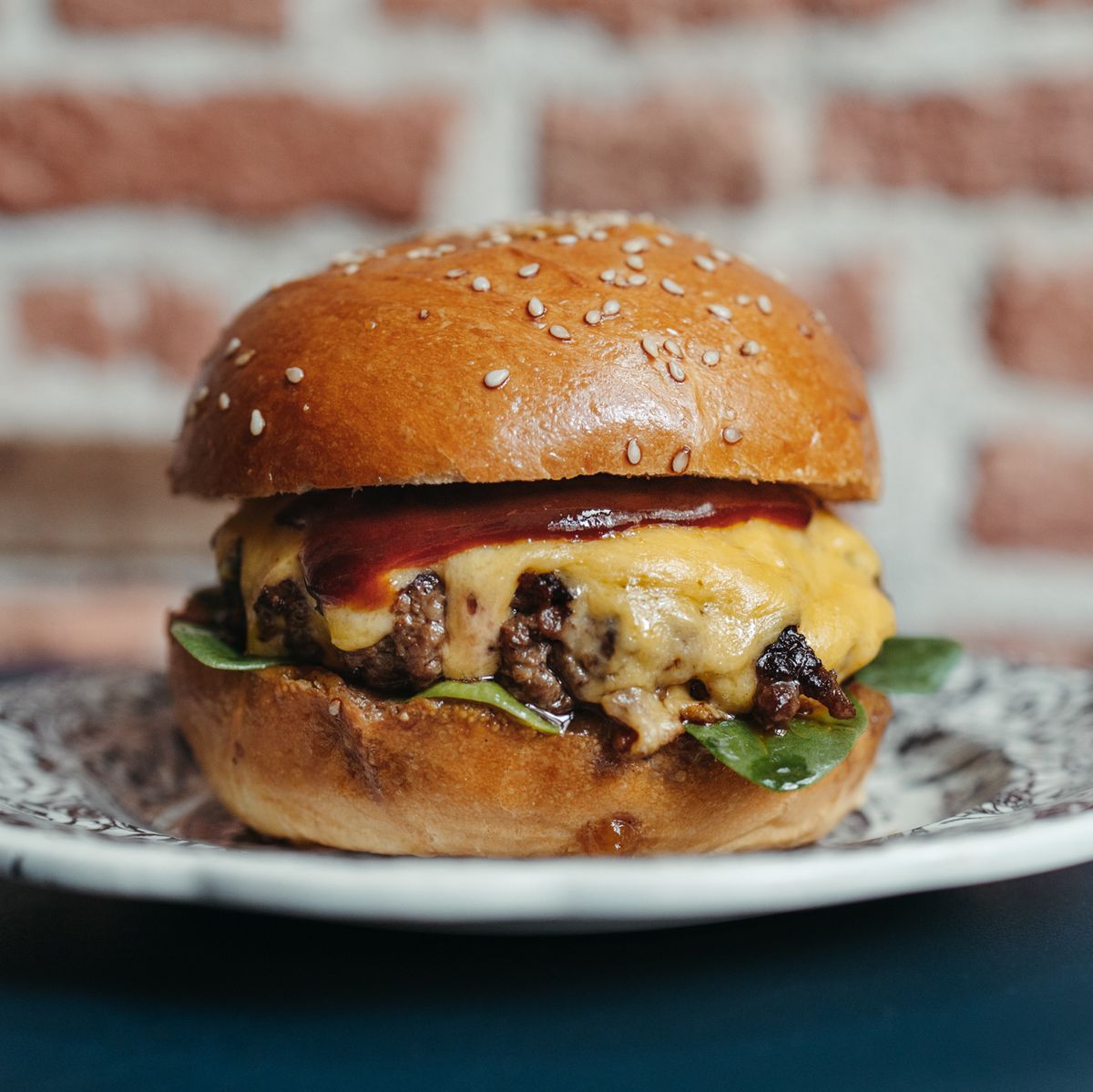 A trim, upright cheeseburger in front of an exposed brick wall.