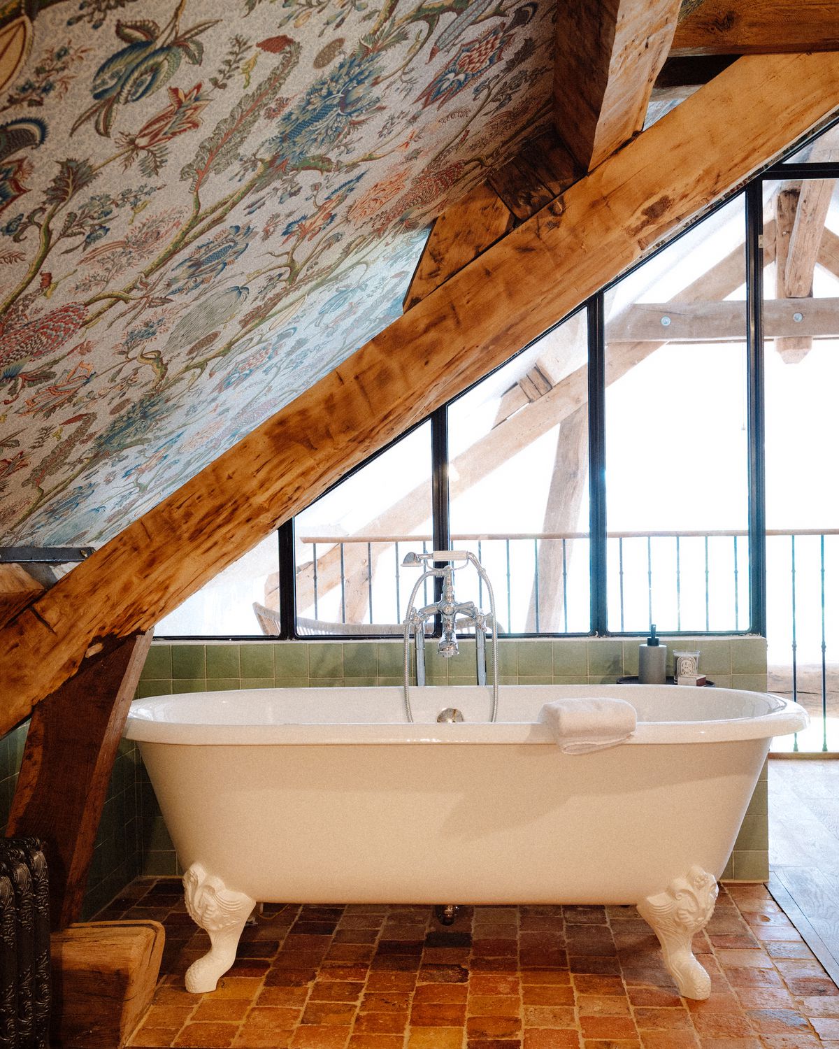 A freestanding bathtub, beneath a slanted ceiling covered with illustrated wallpaper, with a bright patio beyond.