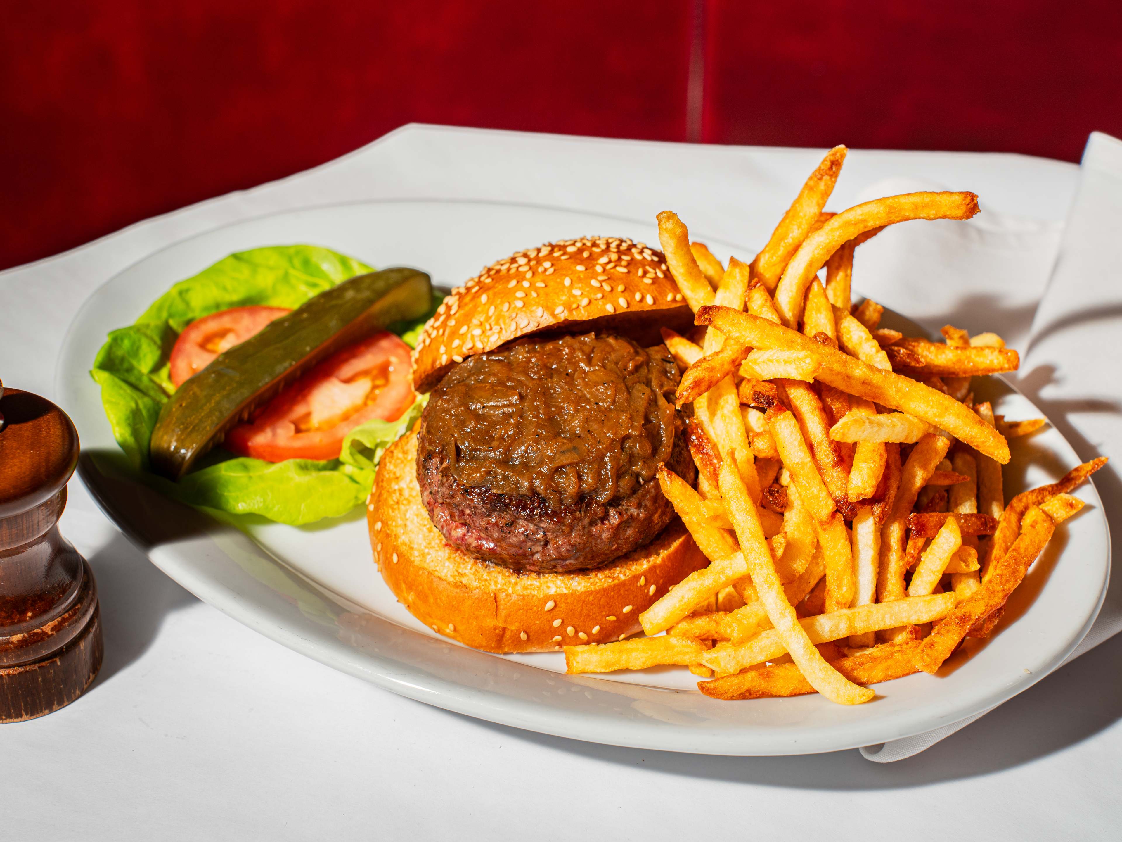 burger patty topped with caramelized onions, with top slightly off the bun. lettuce, tomato and a pickle on one side, and a mess of french fries on the other