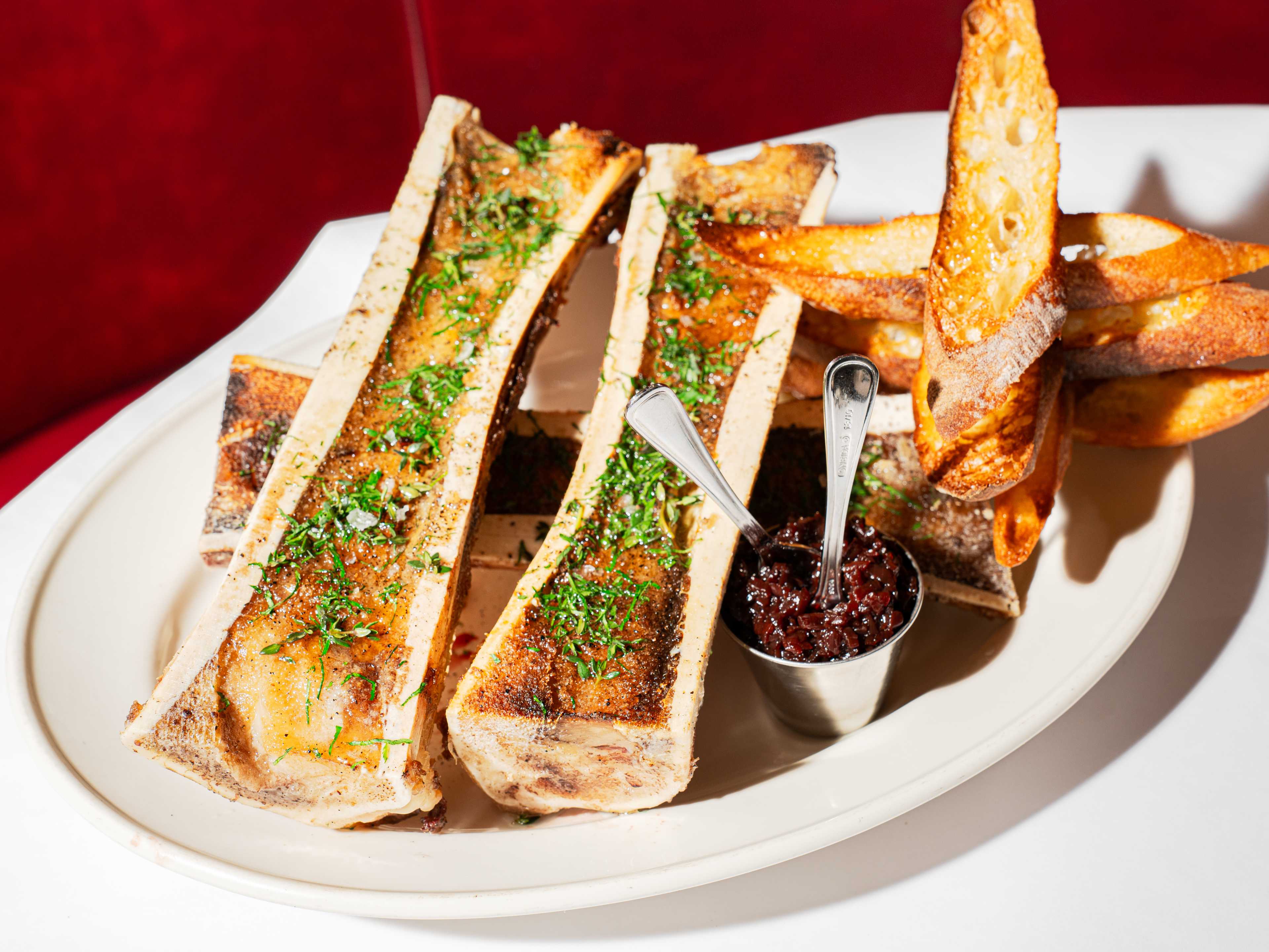 plate with four bone marrow halves sprinkled with herbs, with toasts on the side and a condiment bowl of dark brown shallot jam