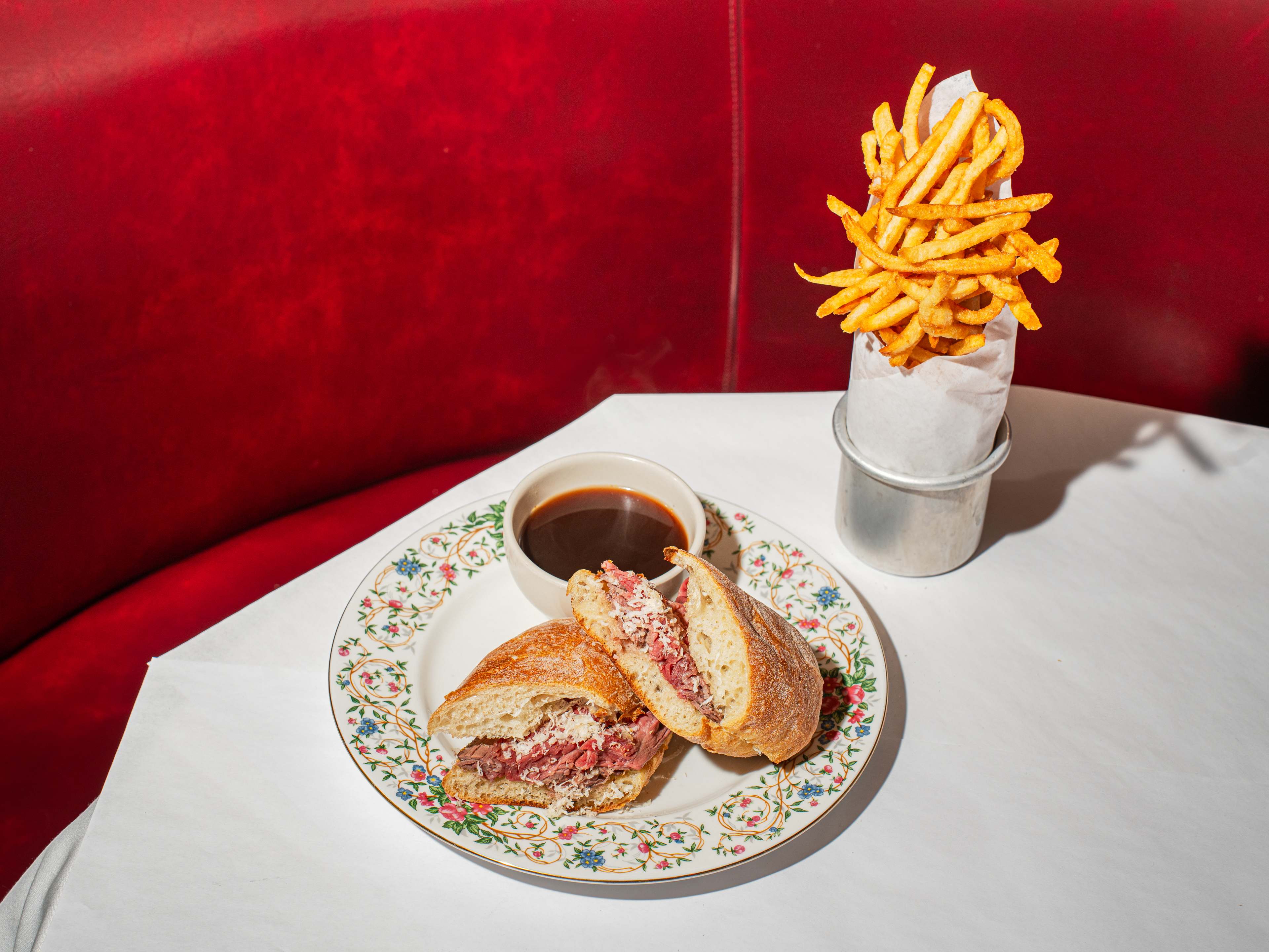 french dip on a floral-rimmed plate with a bowl of au jus. next to  it is a canister of french fries