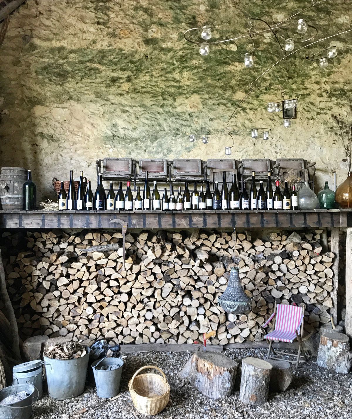 Bottles of wine line a ledge above stacks of cut firewood. 