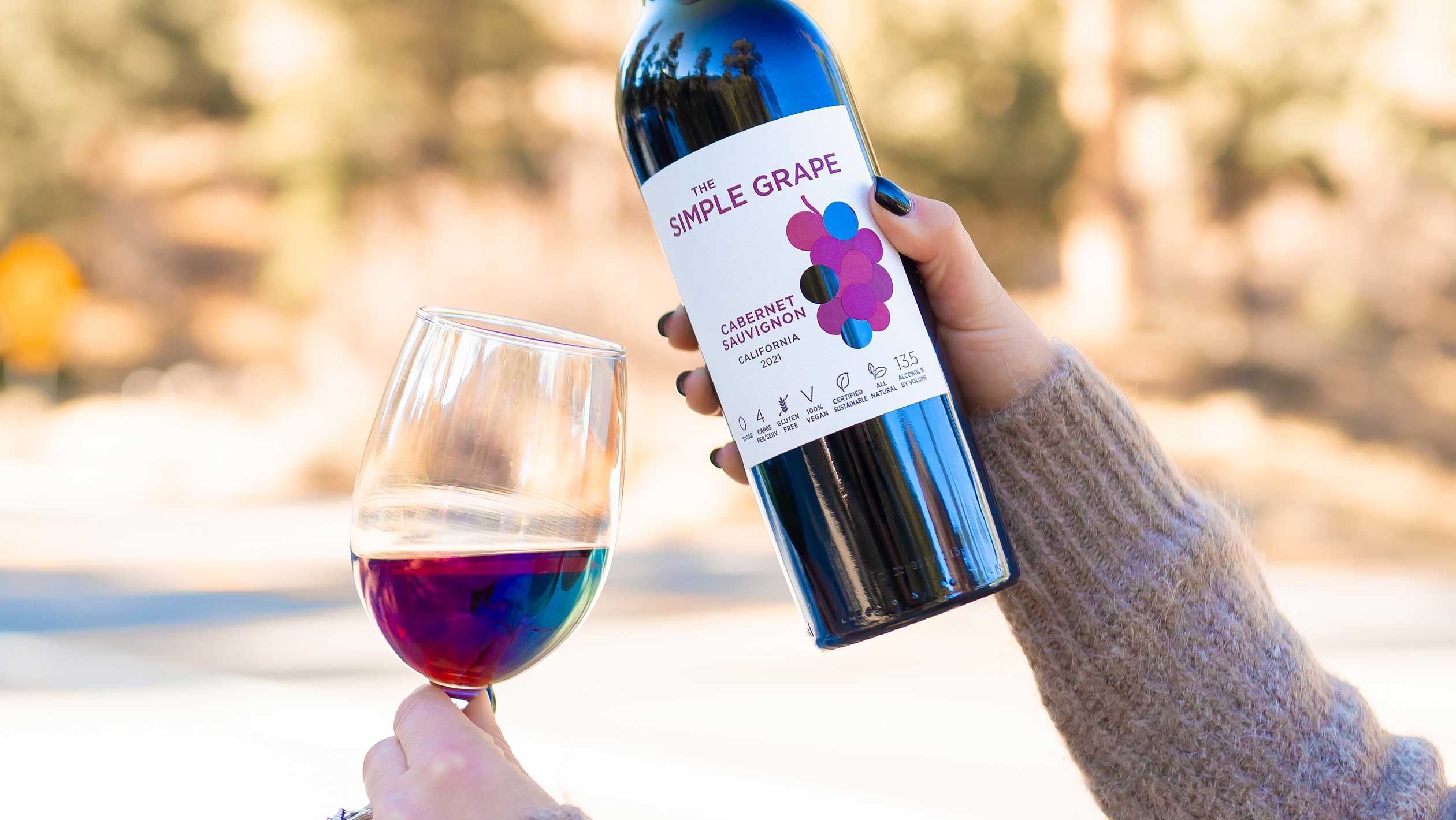 A closeup of hands holding a bottle of Simple Grape and a glass of wine