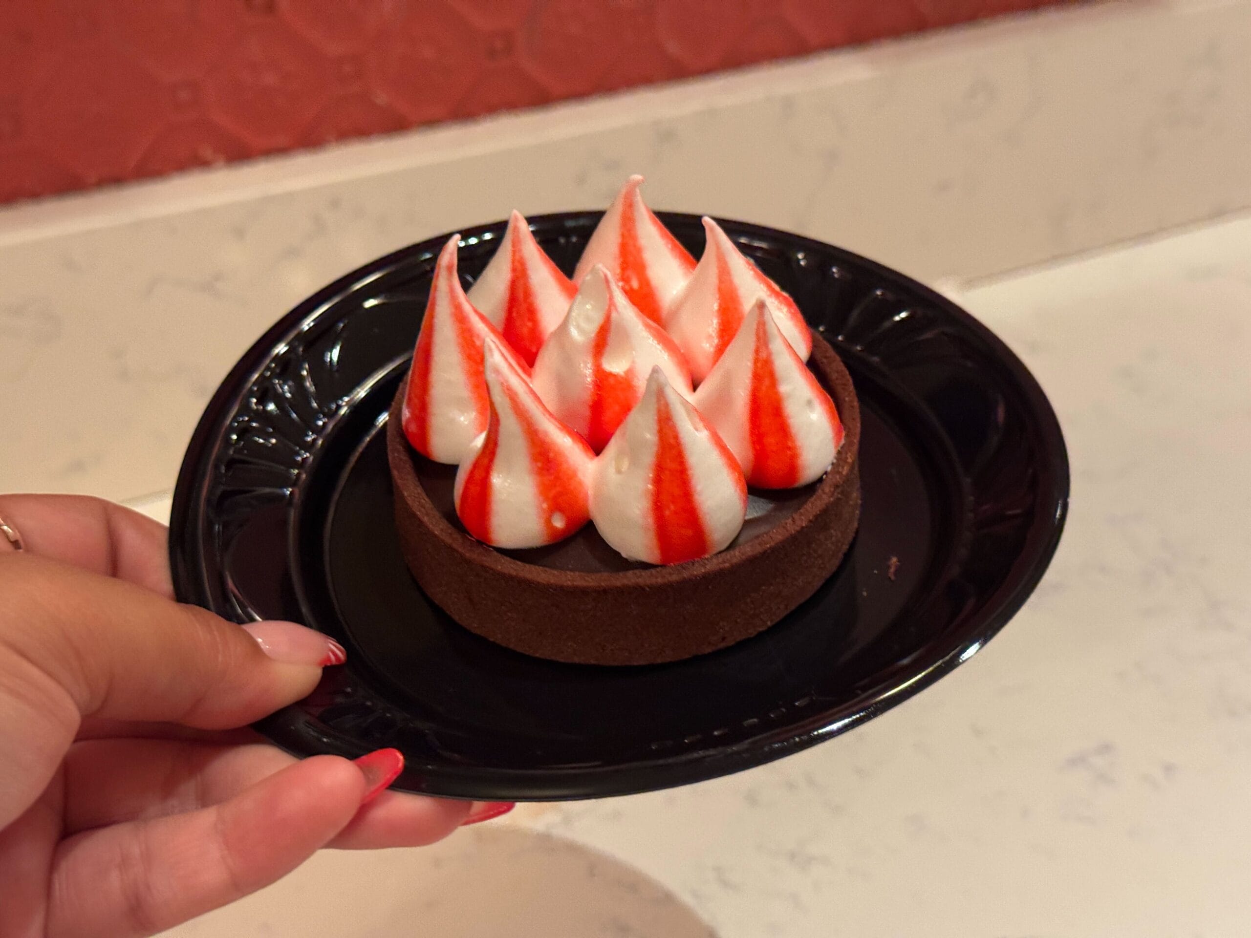 A hand holds a chic black plate at Casey's Corner, presenting a chocolate tart crowned with white meringue kisses elegantly striped with red.