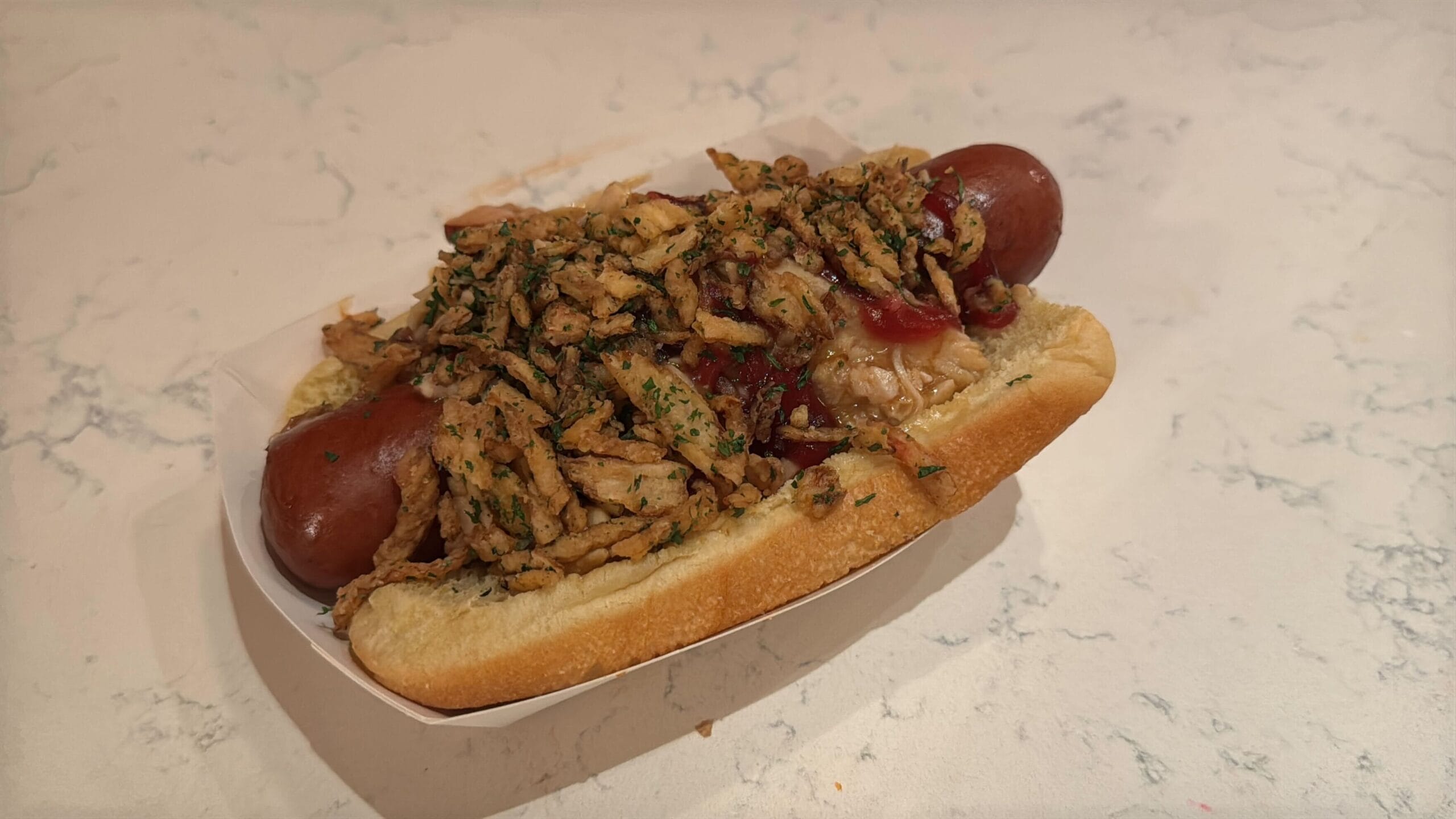 A hot dog from Casey's Corner in a bun topped with crispy fried onions and herbs, elegantly placed on a white paper tray on a light-colored countertop.