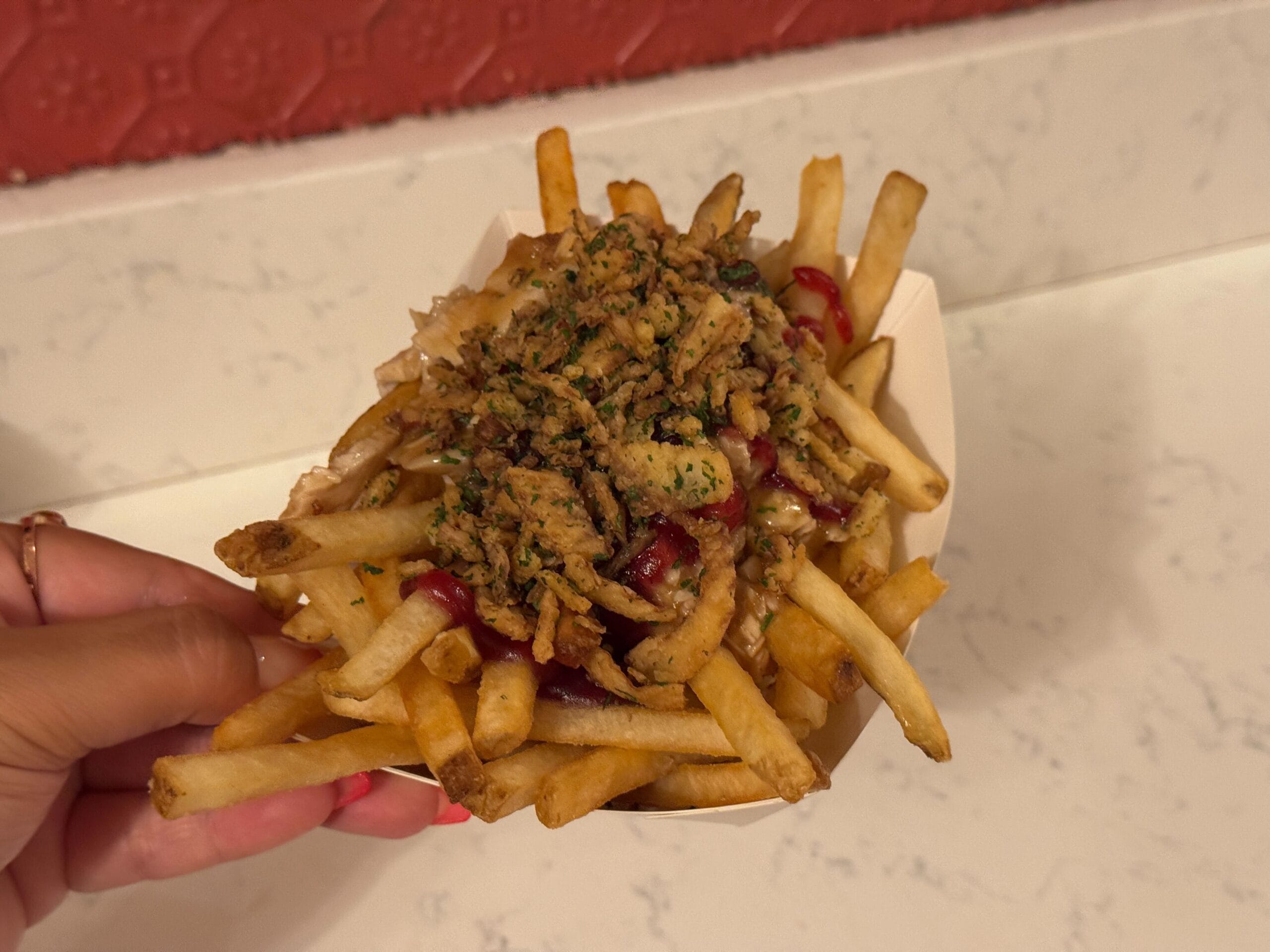 A hand holds a tray of fries topped with sauce and chopped herbs, reminiscent of the delightful offerings at Casey's Corner, against a pristine white countertop.