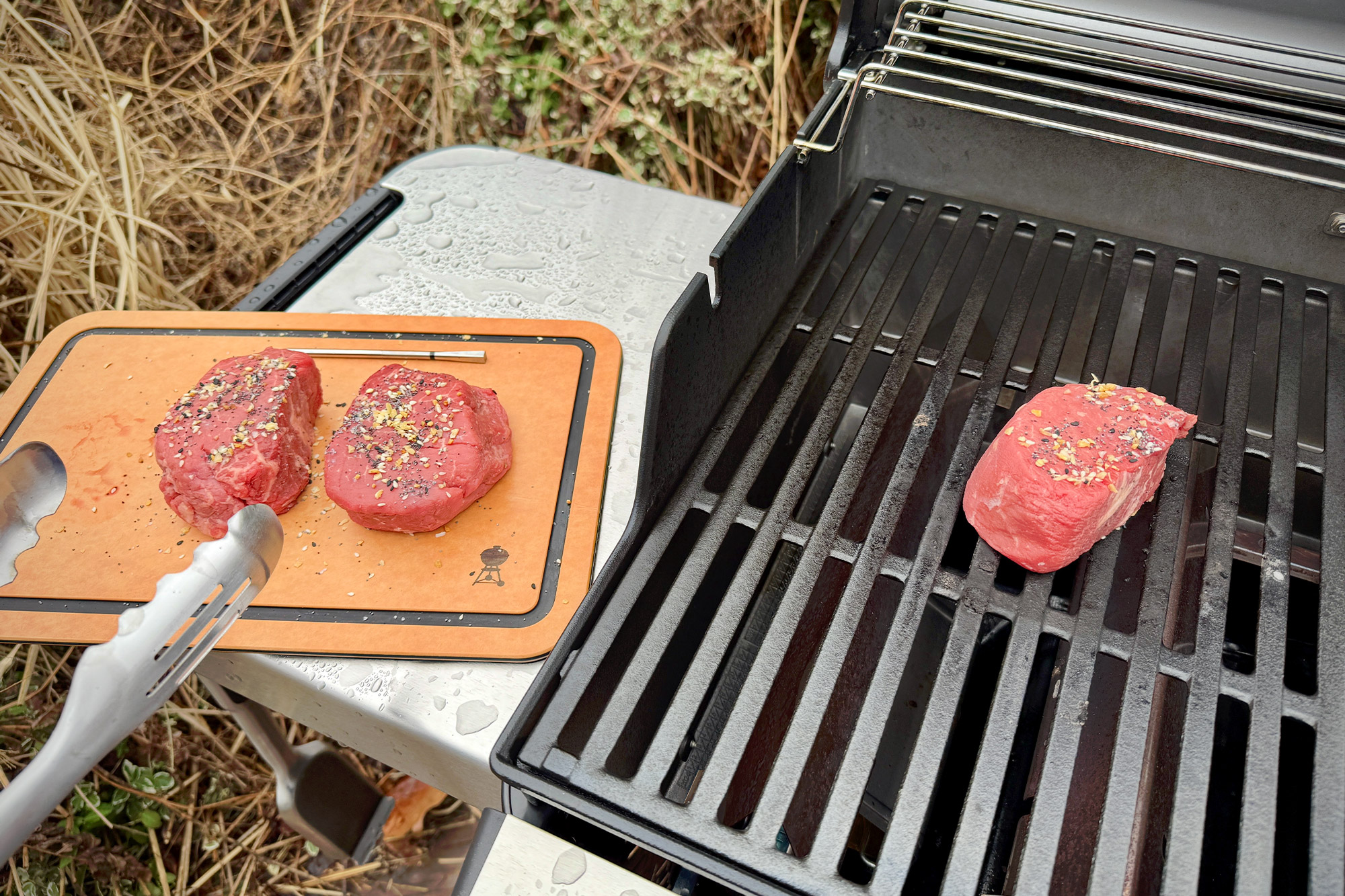 One steak on the Weber Spirit Propane Grill with two seasoned steaks on a cutting board nearby