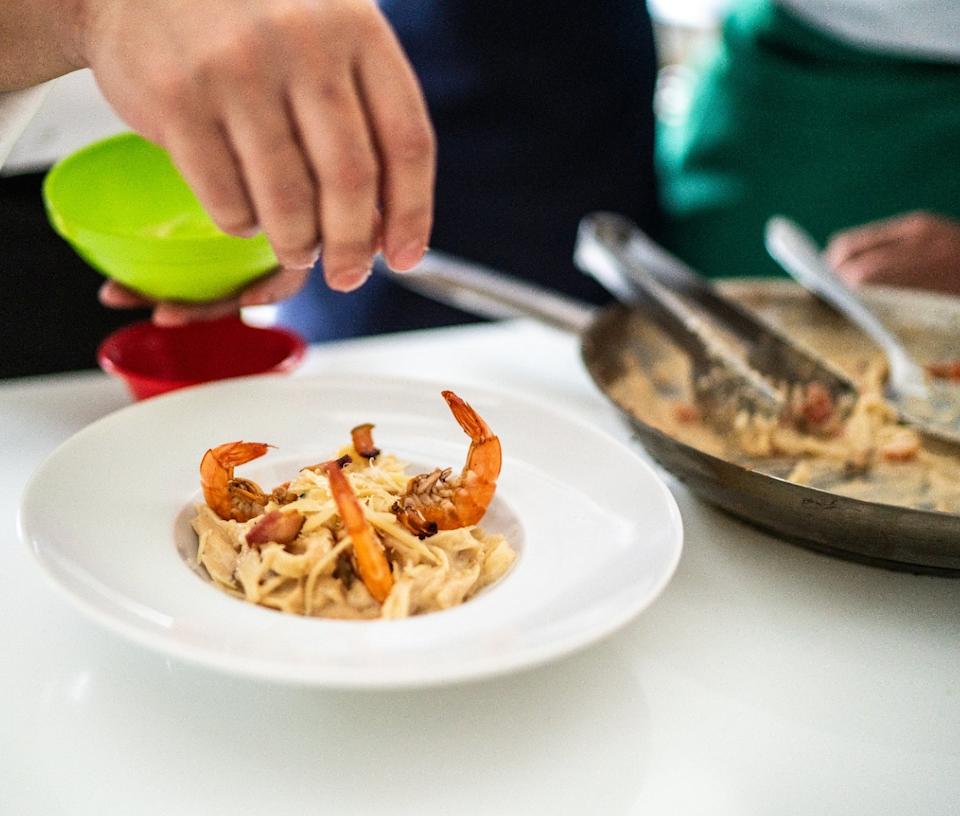 Person garnishing a plate of pasta with shrimp and bacon in a kitchen setting