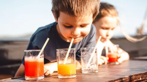 Getty Images A young boy drinks from a glass of juice though a straw while sitting outdoors at a picnic bench in the sun with a friend
