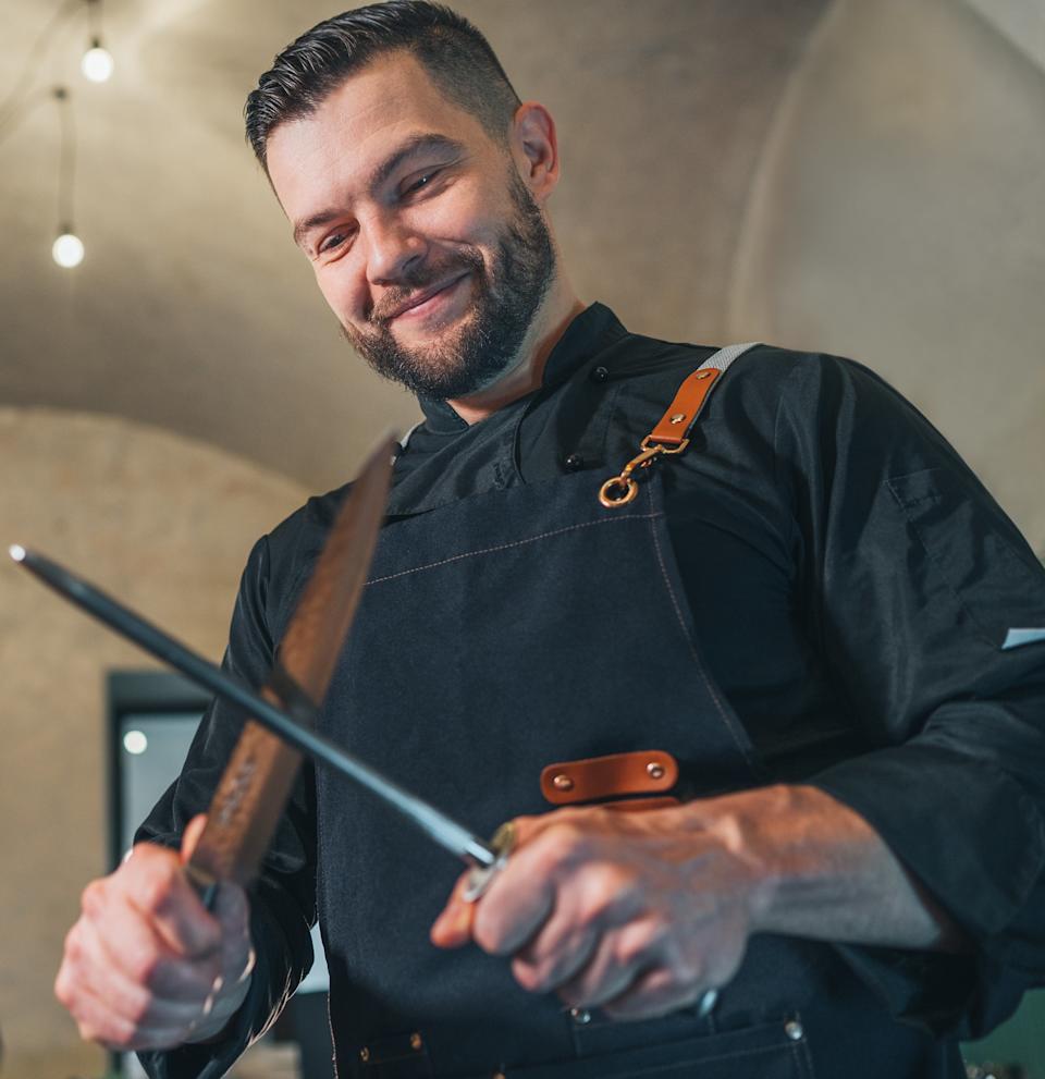 Chef sharpening a knife at a kitchen counter, smiling and dressed in a black chef's jacket with leather details