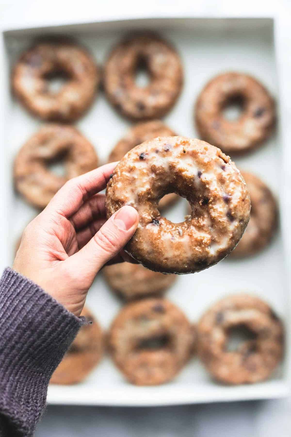 a hand holding a blueberry donut above a box of blueberry donuts.