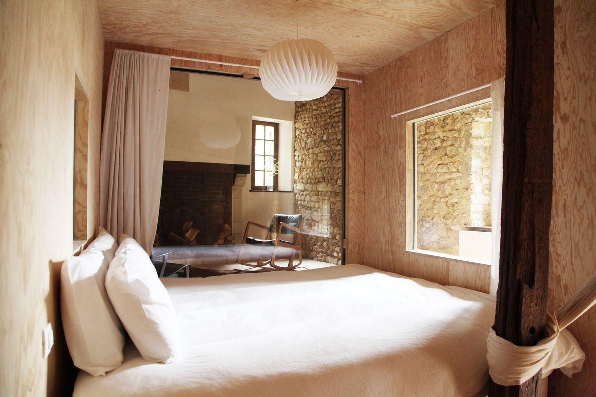 A room at an inn with exposed stone walls, a bed made with fresh linens, a hanging paper lantern, and a large fireplace. 