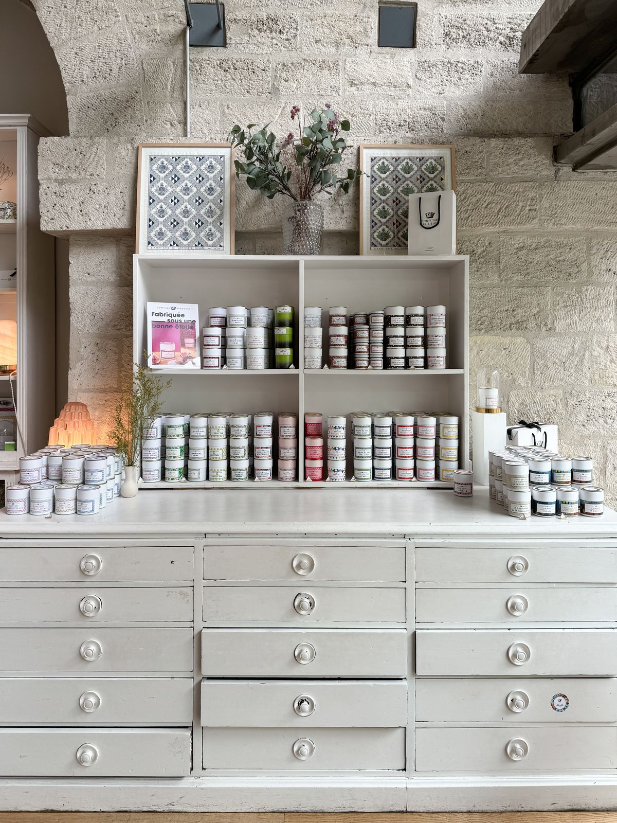 A white cabinet with rows of drawers. On top there are stacks and shelves of jam jars.